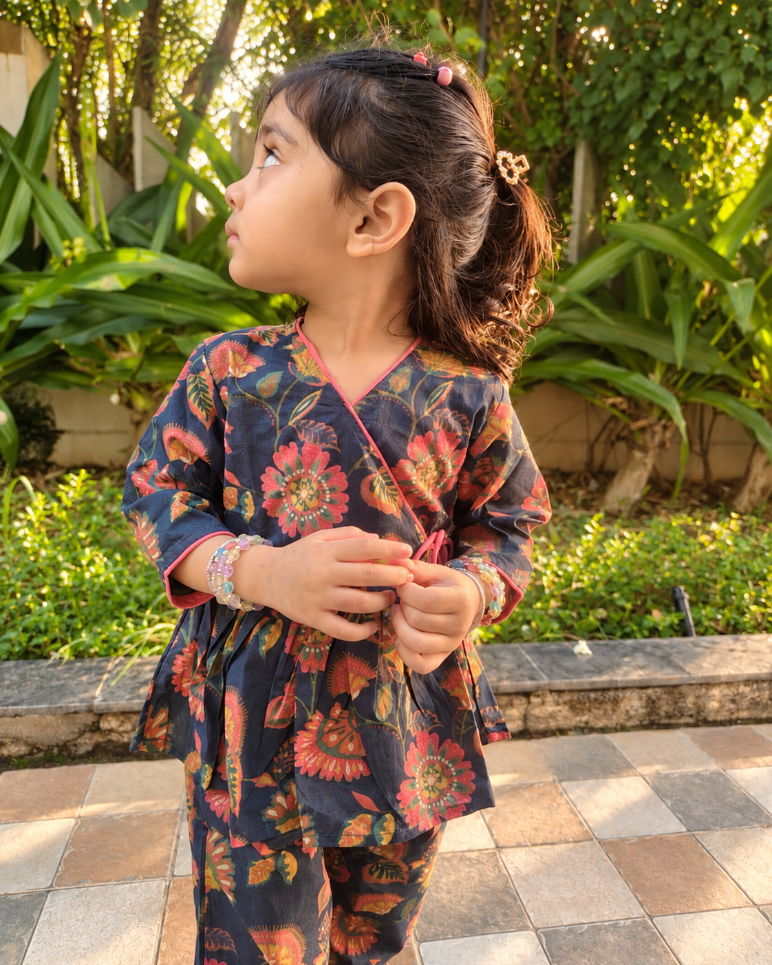A young girl wearing a dark navy floral Angrakha top and matching pants, from Aruvo stands outdoors in a garden setting. The outfit features a vibrant multicolor floral pattern with red, orange, and yellow flowers. 