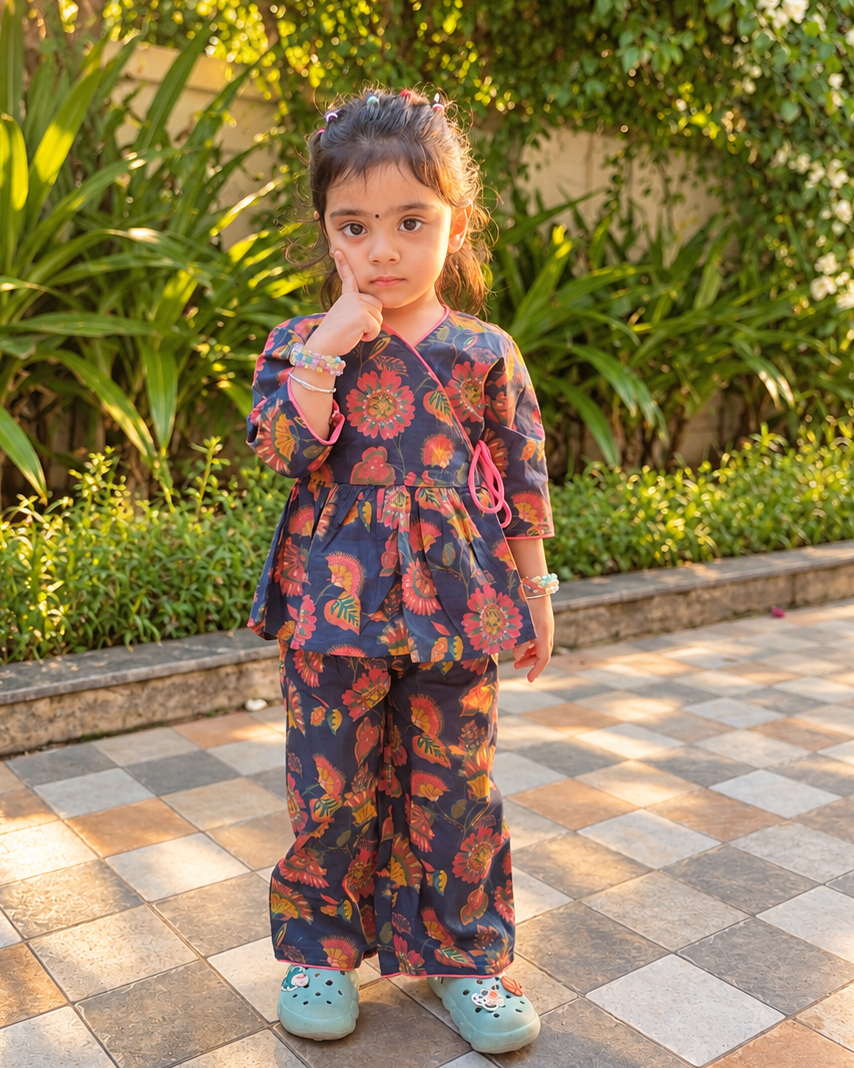 A young girl stands on a tiled patio in a garden setting, wearing a matching two-piece outfit with a dark blue base and a vibrant orange, red, and yellow floral pattern. She is wearing light blue Croc-style shoes.
