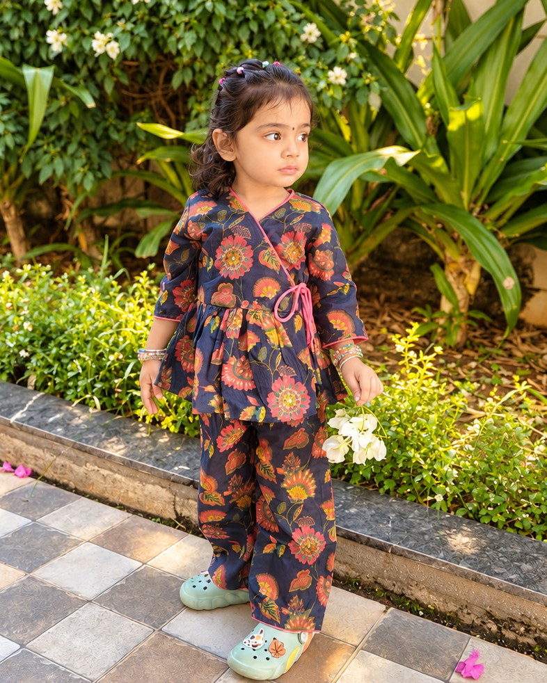 A young girl stands on a tiled patio in a garden setting, wearing a dark navy two-piece outfit with a vibrant orange, yellow, and red floral pattern, paired with light green Crocs.