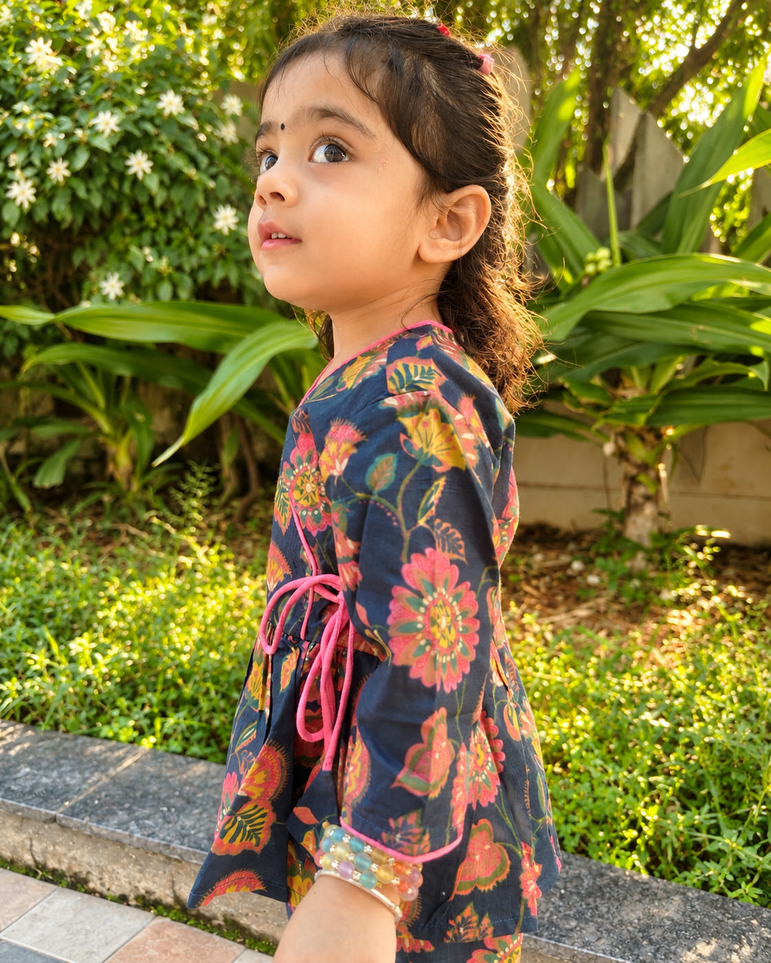 A young girl wearing a navy blue floral printed kurta with pink and orange details, standing outdoors in a garden setting.
