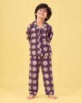A young boy wearing a purple pajama set with a white circular pattern, standing against a solid pale yellow background.