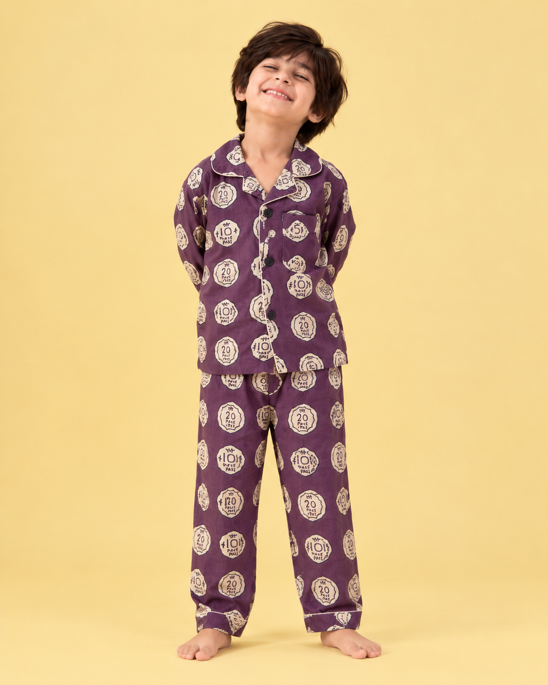 A young boy wearing a purple pajama set with a white circular pattern, standing against a solid pale yellow background.