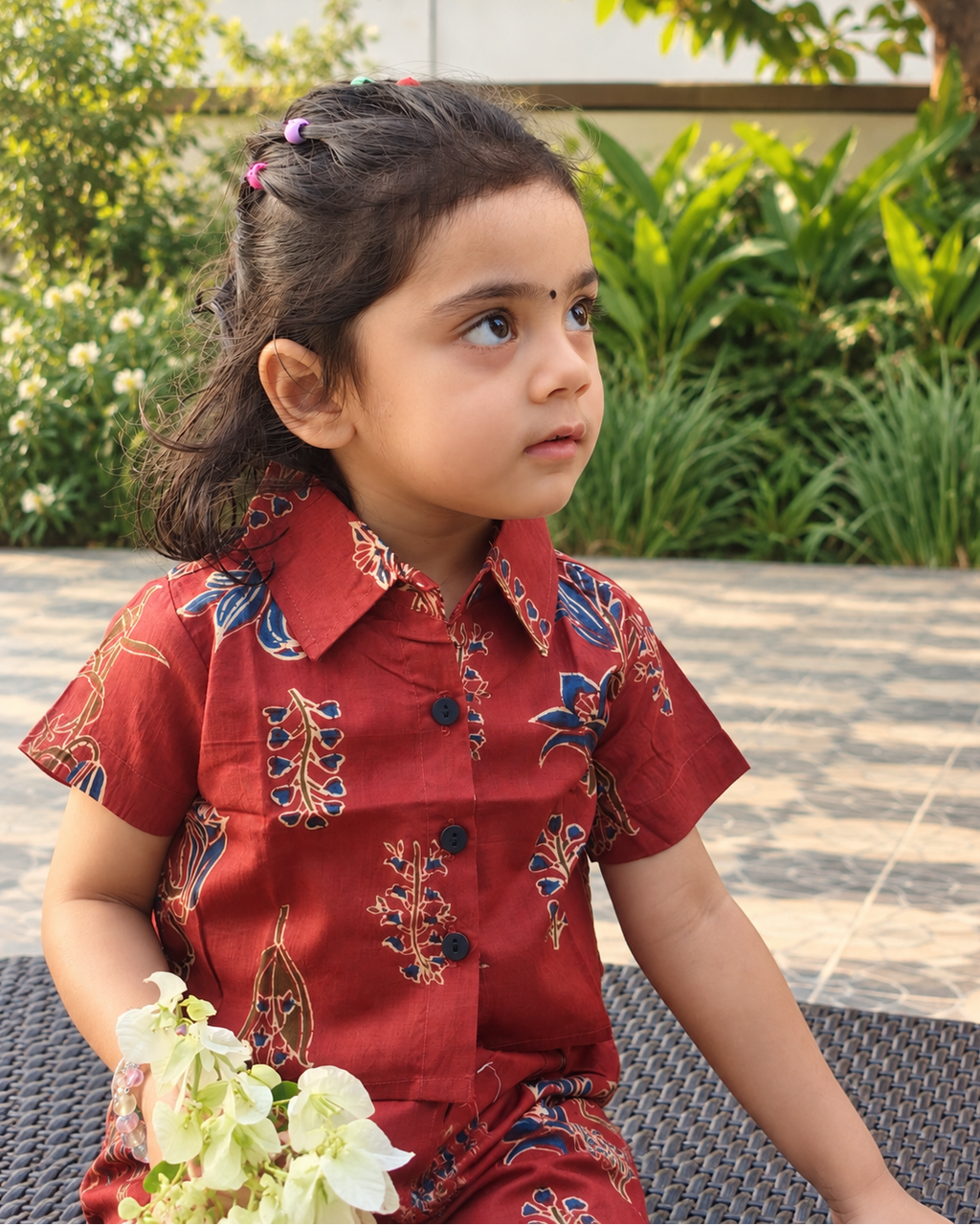 A young girl with dark hair in a ponytail sits outdoors wearing a rust short-sleeve shirt in floral patterns. She is holding a small bouquet of white flowers.