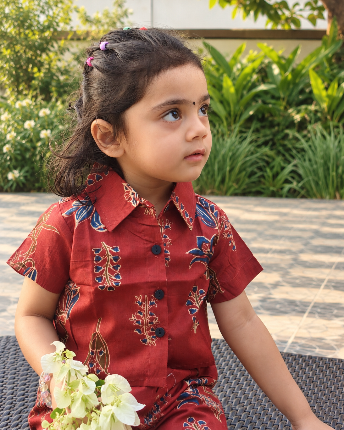 A young girl with dark hair in a ponytail sits outdoors wearing a rust short-sleeve shirt in floral patterns. She is holding a small bouquet of white flowers.