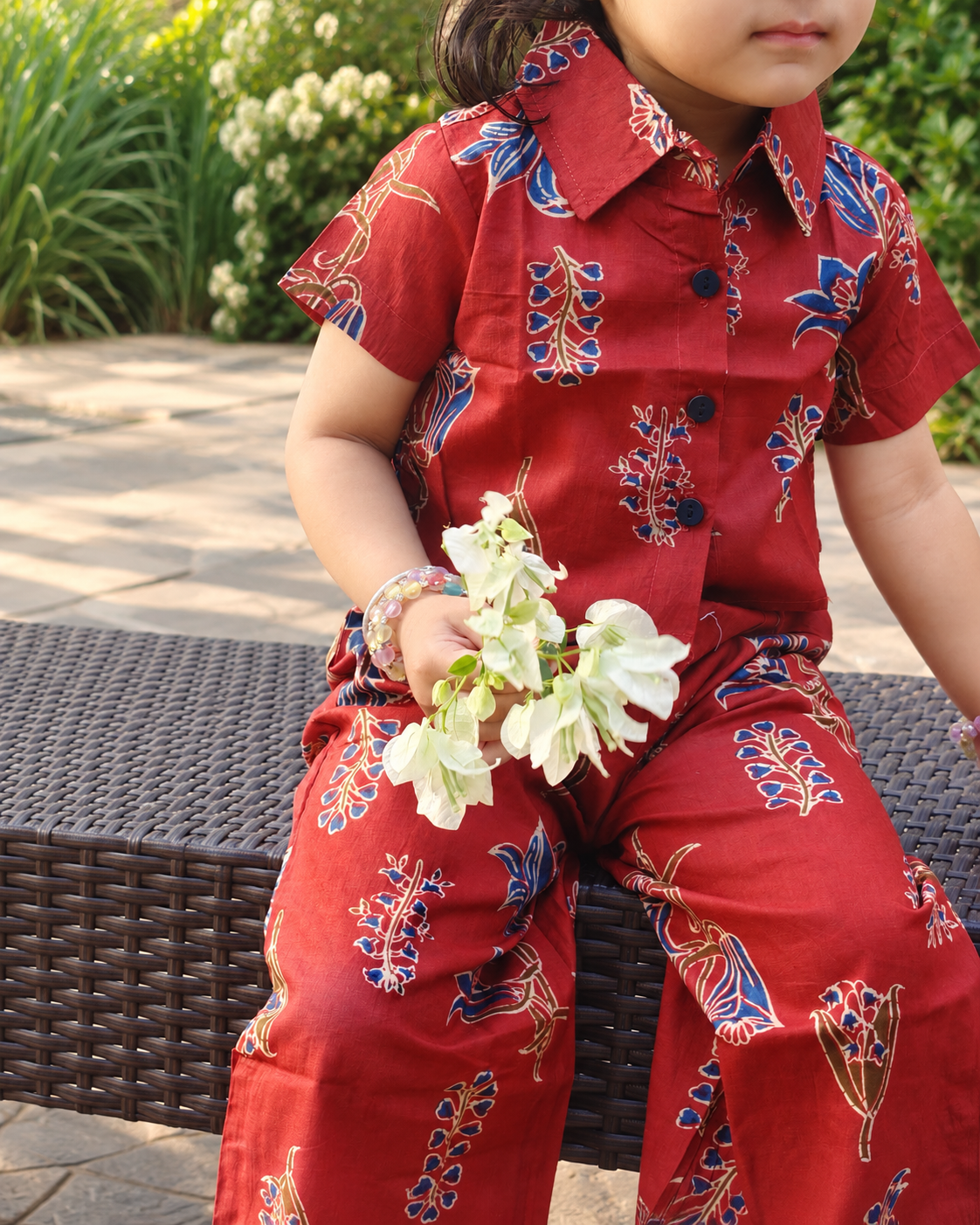 A child wearing a rust short-sleeve button-up shirt and matching pants with a blue and gold batik-style floral pattern, sitting outdoors on a dark woven bench and holding white flowers.