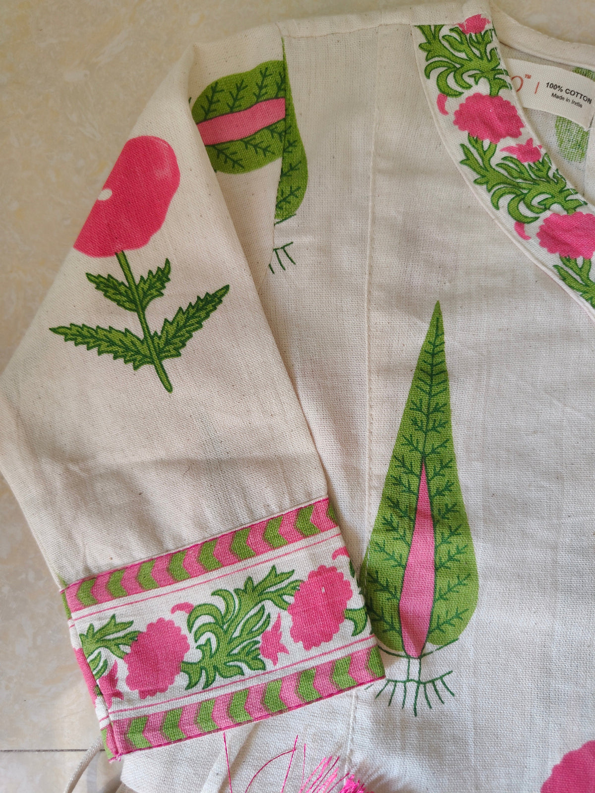A close-up of a white or off-white fabric garment, likely a kurta or dress, featuring hand-block printed motifs of pink flowers and green leaves, with a decorative pink and green striped border.