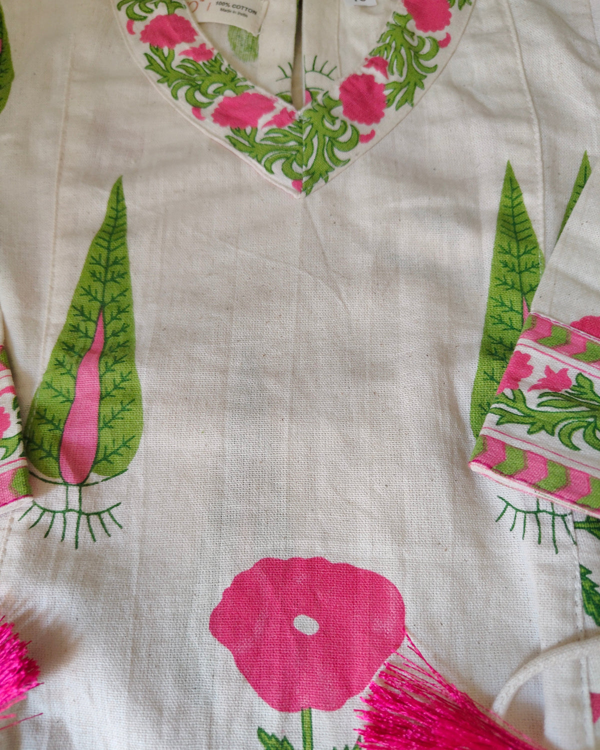 A close-up of a white fabric garment featuring a colorful print of pink flowers, green leaves, and tassels.