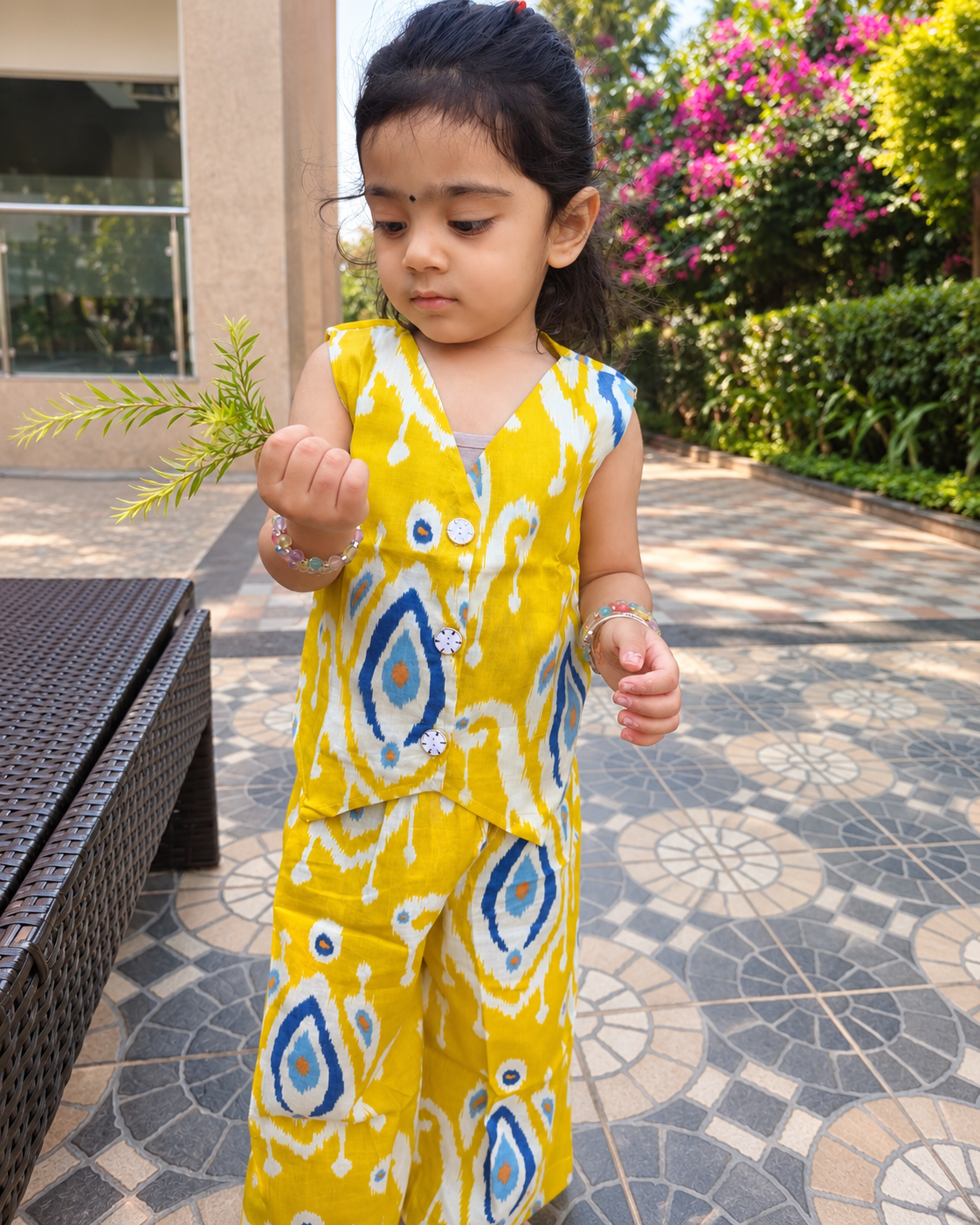 A toddler girl wearing a bright yellow sleeveless waistcoat pattern top and matching pant set with a blue and white ikat pattern, standing on a patterned outdoor patio.