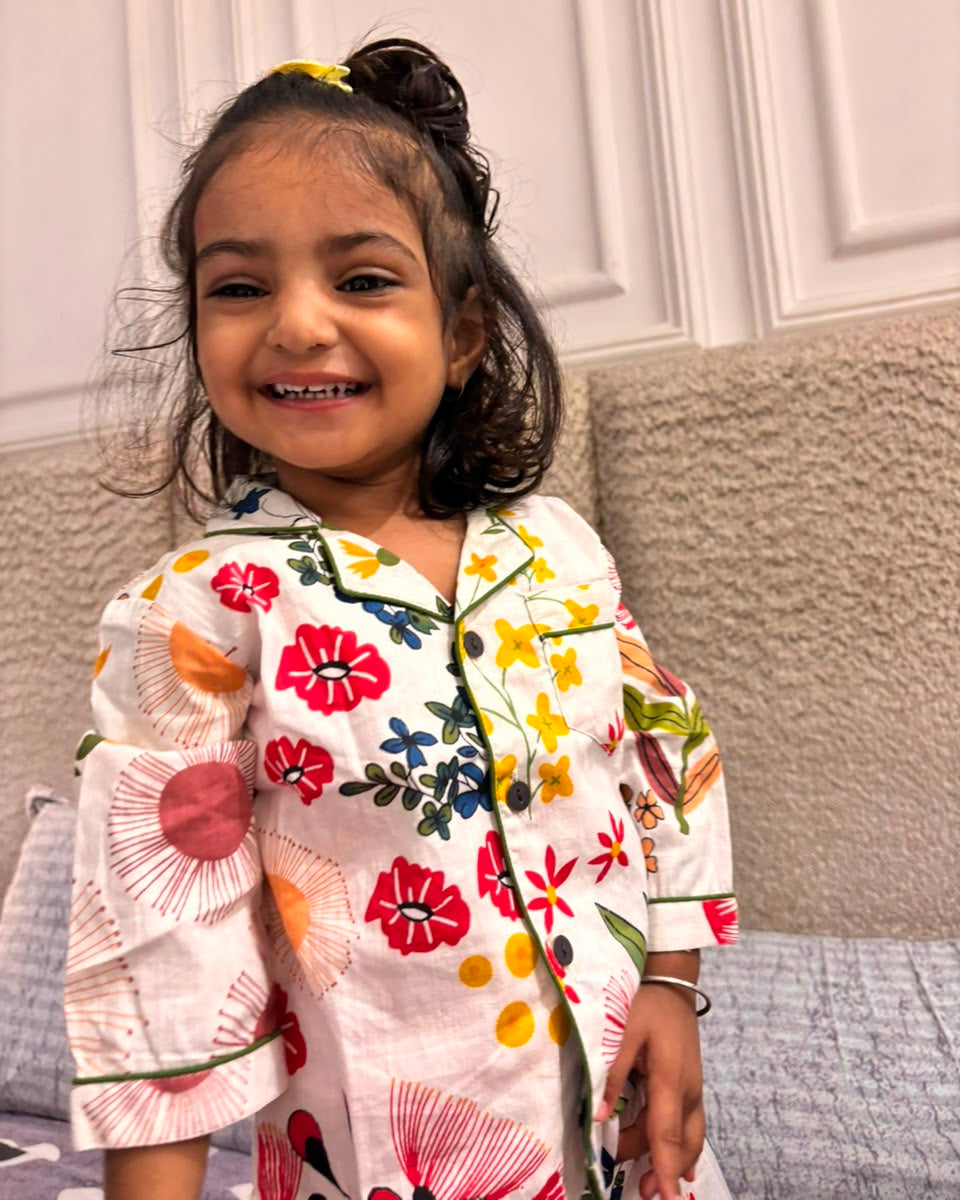 A toddler is wearing a loral printed full sleeve nightsuit set in white color from brand Aruvo, standing on a bed smiling 